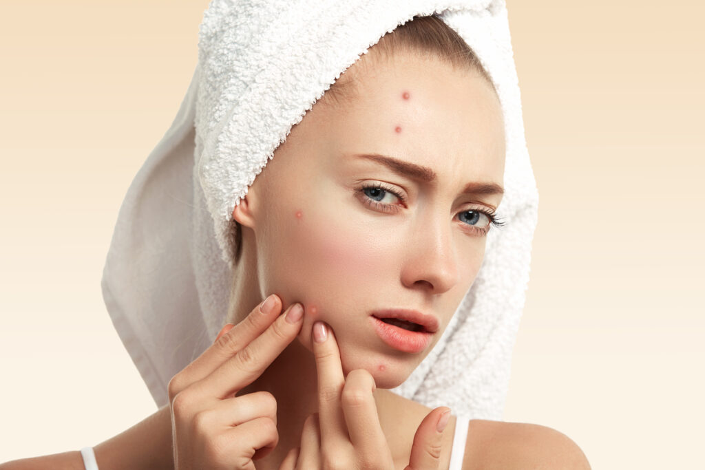 Close up isolated portrait of young woman squeezing pimple on her cheek in the bathroom. Cropped view of Caucasian female having skin problem, against blue wall background. Beauty and spa concept