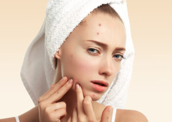 Close up isolated portrait of young woman squeezing pimple on her cheek in the bathroom. Cropped view of Caucasian female having skin problem, against blue wall background. Beauty and spa concept